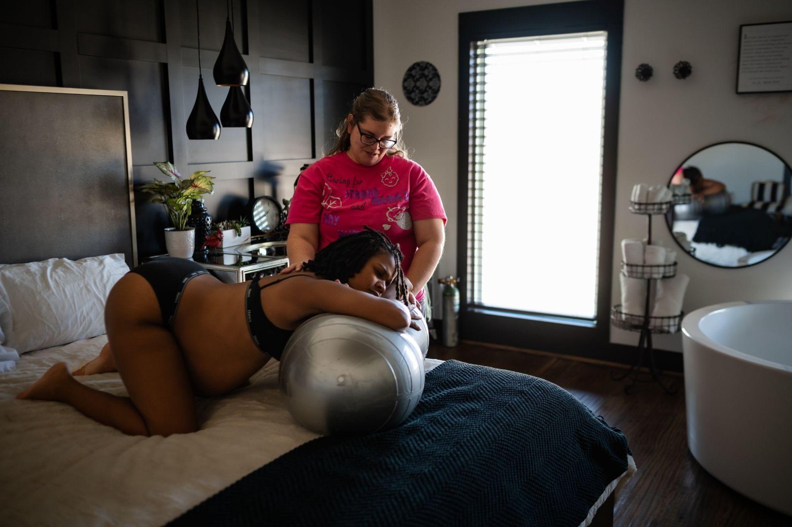 A pregnant woman leaning over a bed while a woman in a pink shirt holds her hands